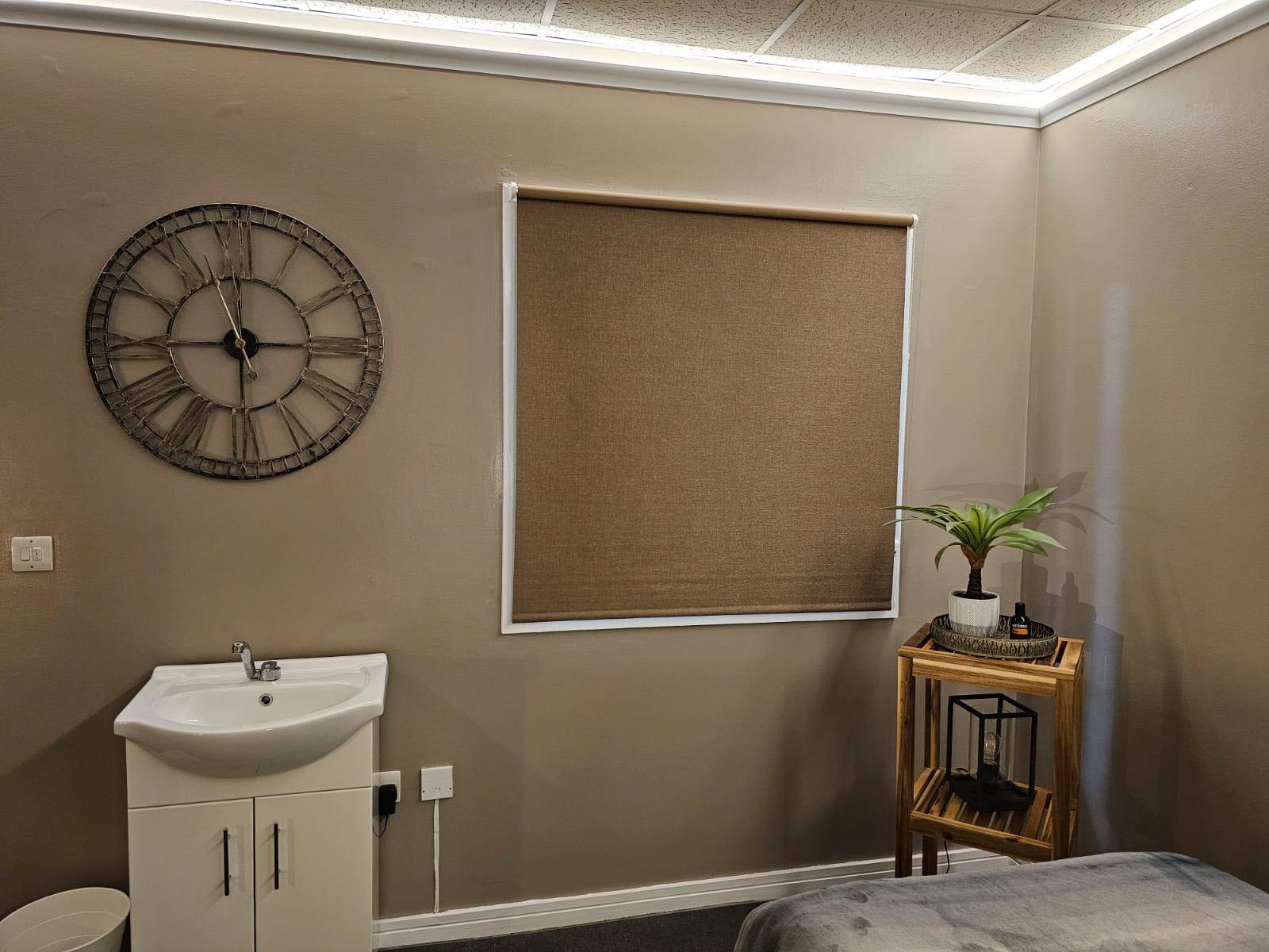The other side of the room: a large clock, a woven blind, the basin unit and a plant on a wooden shelf