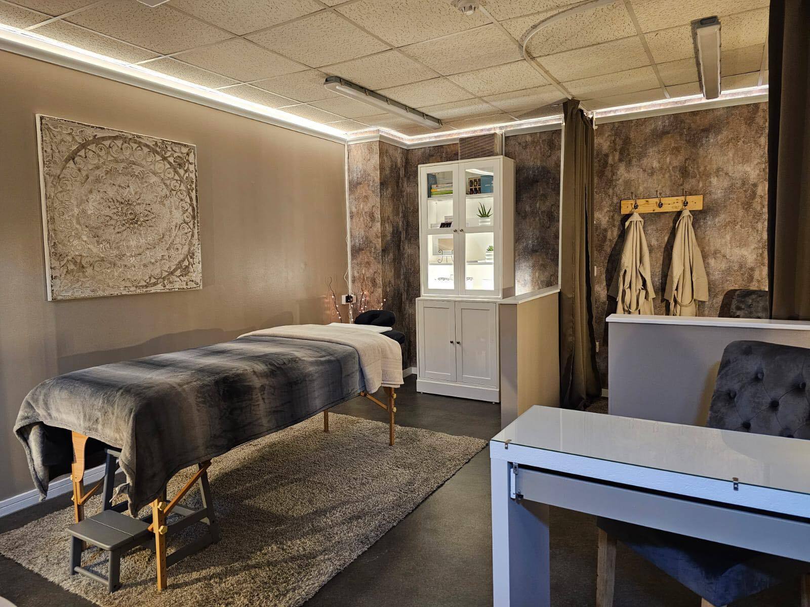 The treatment room: massage couch with a striped throw, bronze textured walls, a mandala artwork, a glass cabinet and robes on hooks